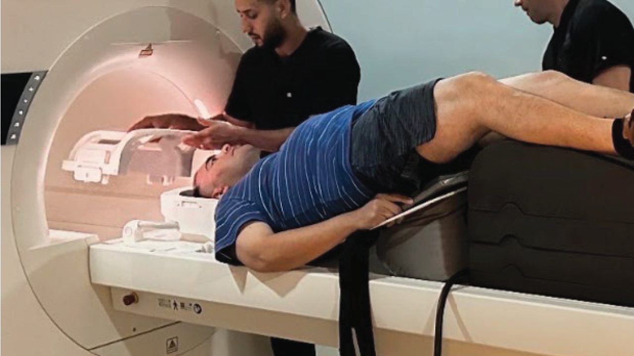Technologist seating a head coil; patient on the table with the MRI bore behind